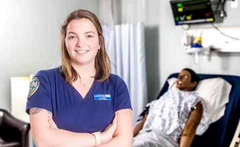 Portrait of Anne Carrigg, a nursing student, in UNE's simulation lab. A patient simulator is behind her.